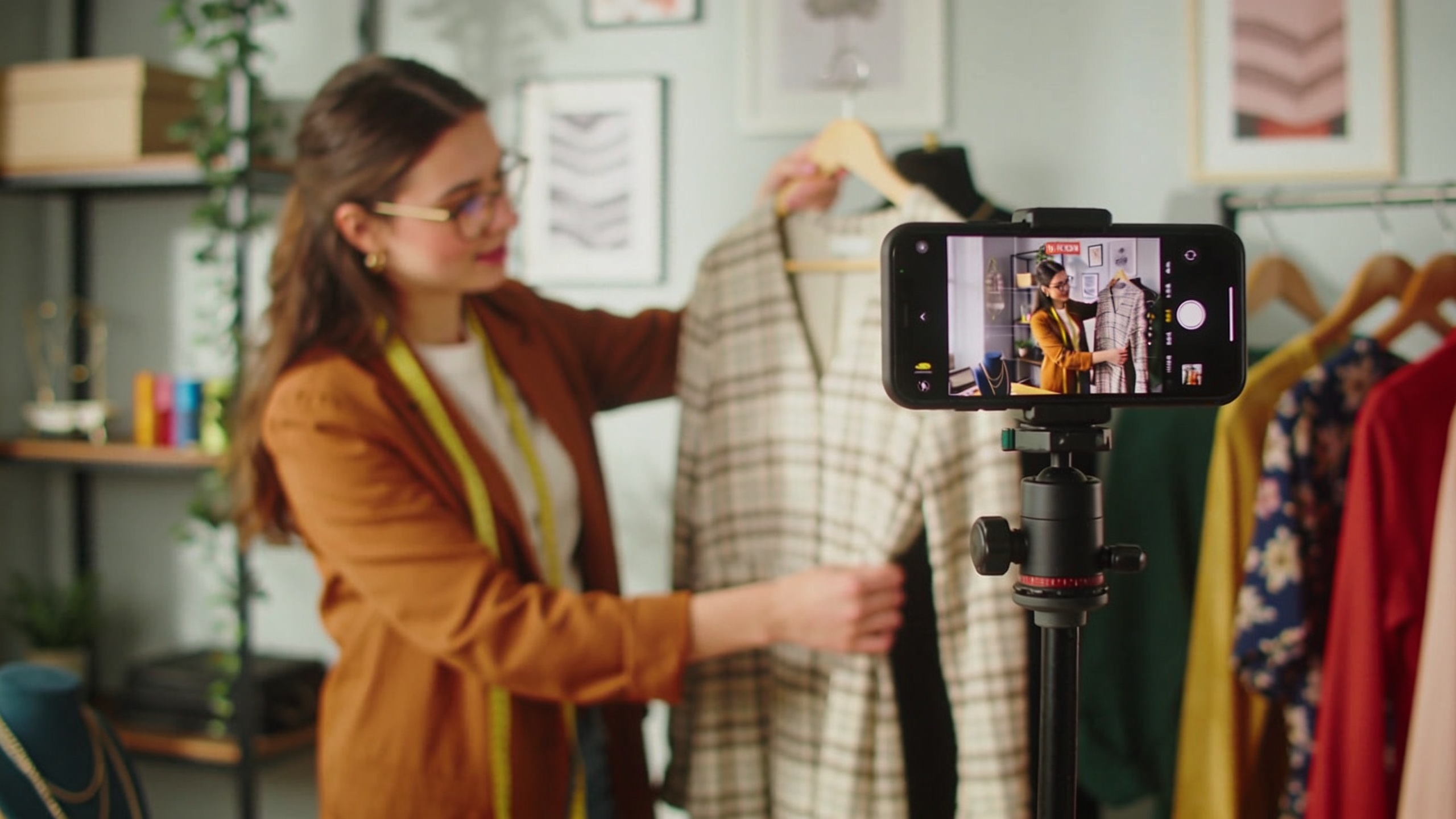 Jeune femme propriétaire de boutique filmant un tutoriel mode avec son smartphone, présentant un vêtement de sa collection, dans une ambiance chaleureuse et authentique, style vidéo Instagram Reel, lumière naturelle.