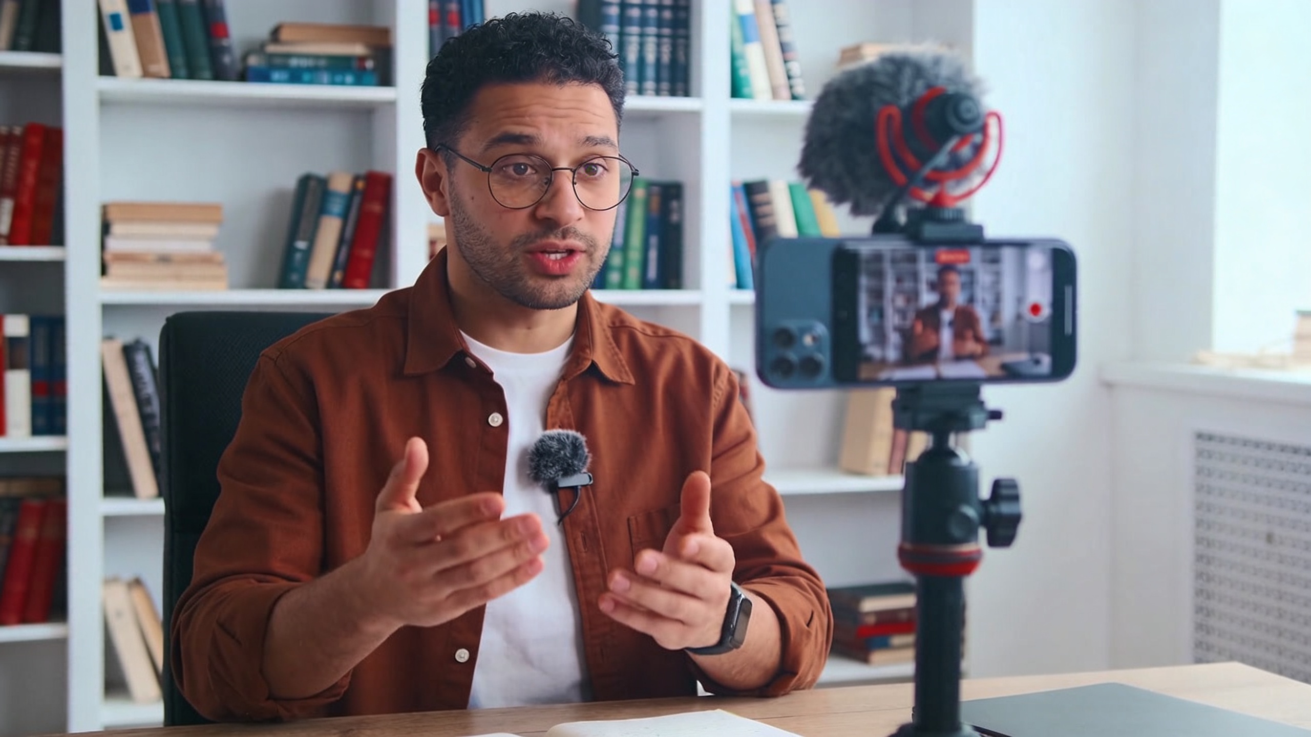 Coach en train de filmer un Reel Instagram avec son smartphone, expliquant un concept de développement personnel. Un micro cravate est visible, et l'arrière-plan est un bureau moderne avec des livres. Lumière douce, style vlog professionnel, couleurs naturelles et inspirantes.