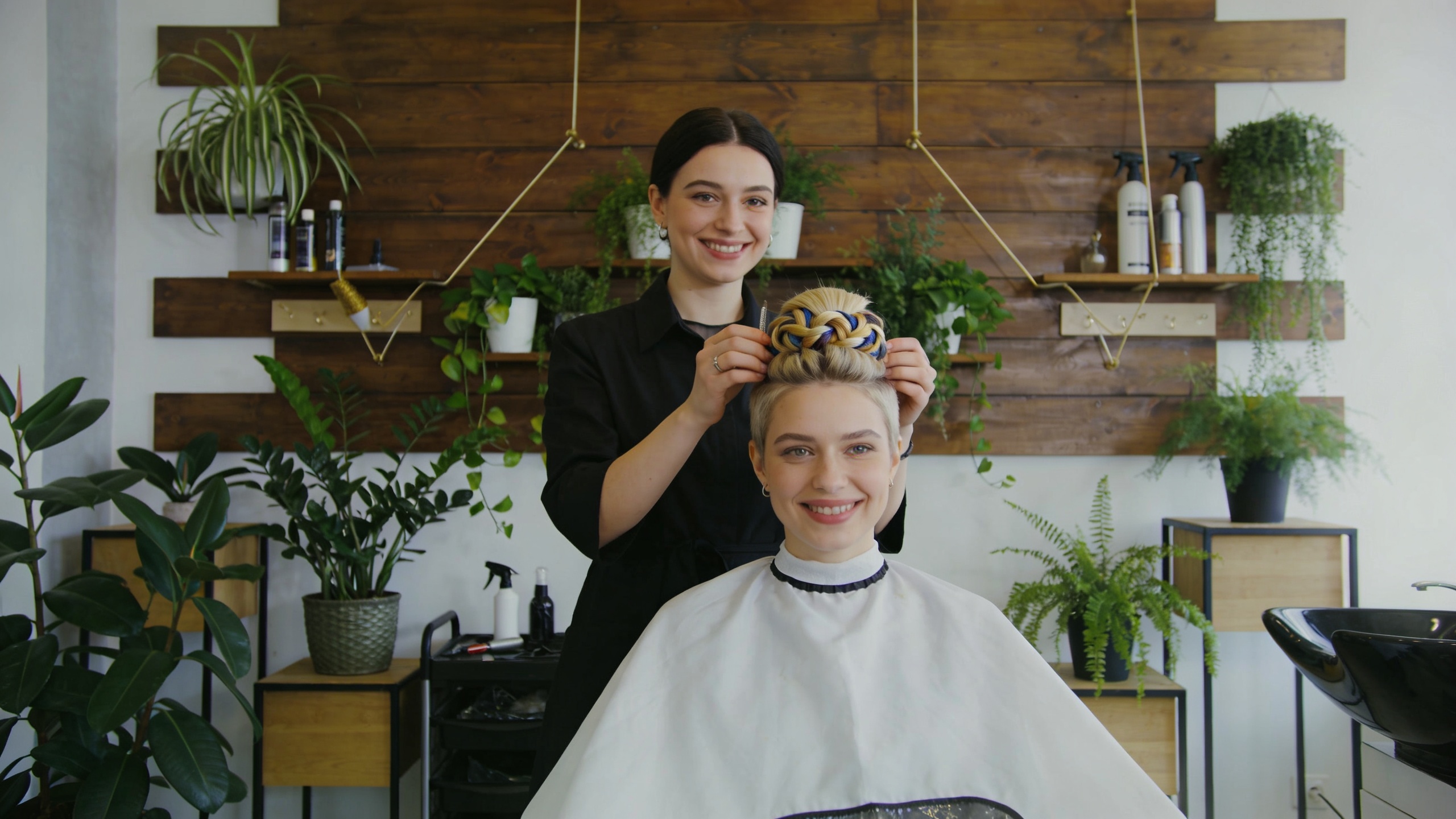 Coiffeuse montrant fièrement une coupe de cheveux créative et originale à une cliente ravie, dans un salon de coiffure design avec des éléments végétaux, lumière douce et naturelle, style photo éditorial magazine, composition cinématique.