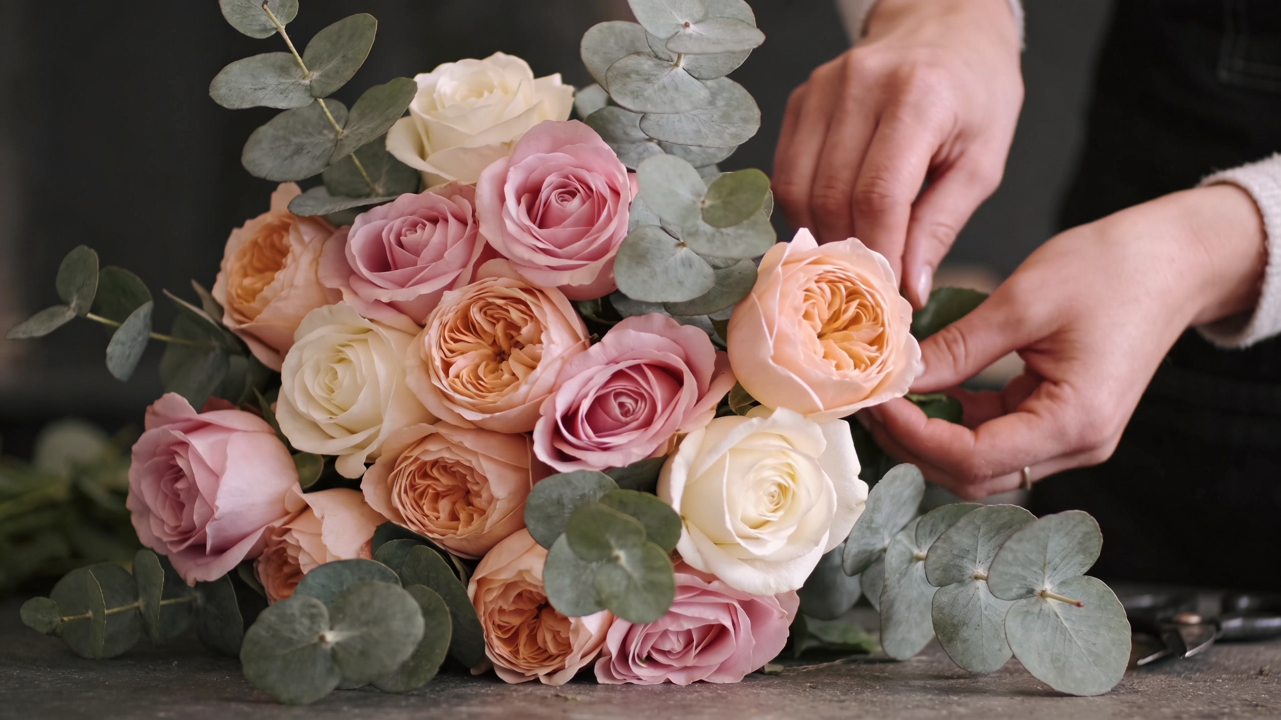 Gros plan détaillé sur des mains de fleuriste composant un bouquet de roses pastel et d'eucalyptus, lumière naturelle douce, style photo artistique et éditorial, mettant en valeur la texture des pétales.