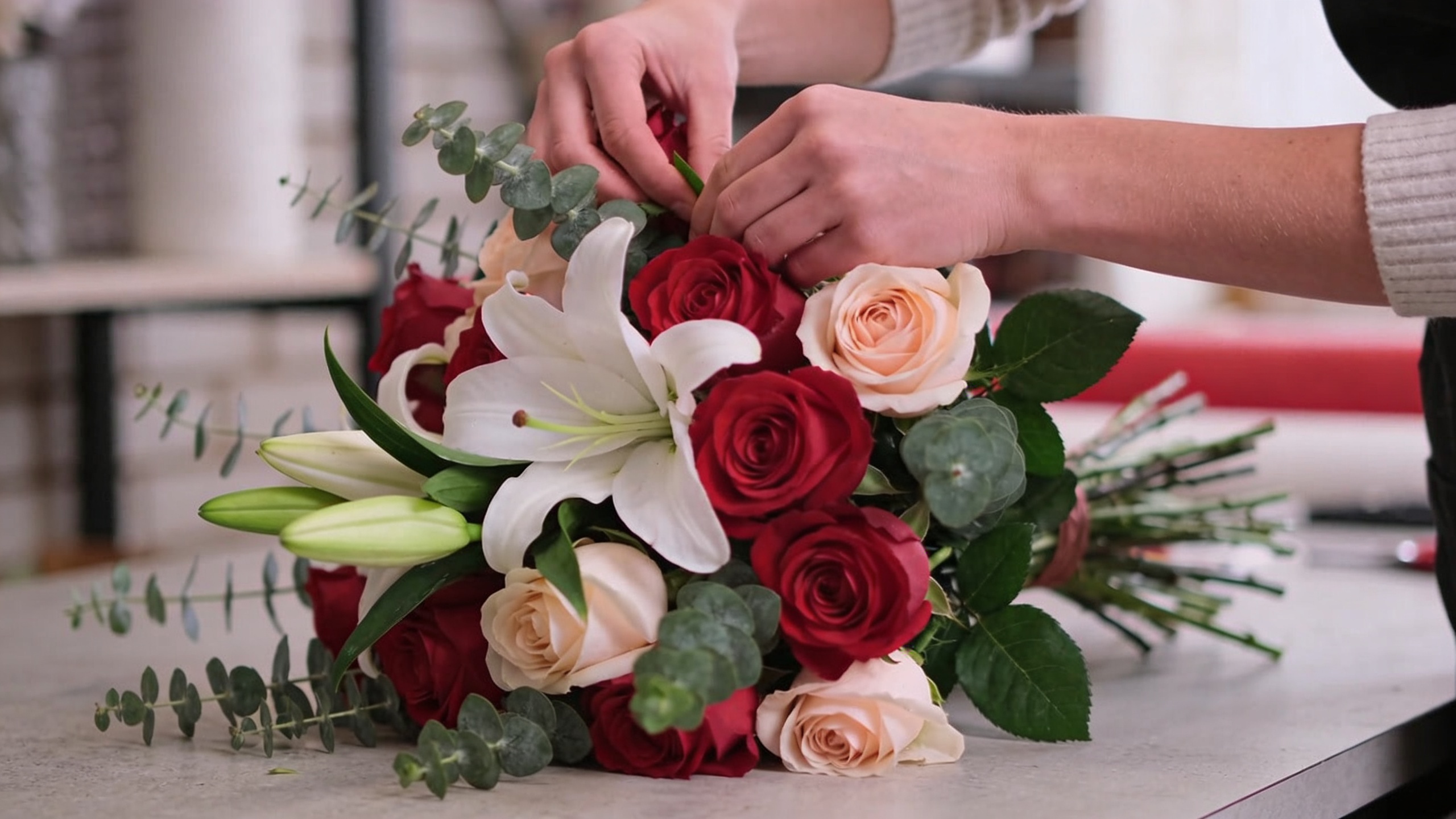 Gros plan sur les mains d'un fleuriste expert, composant un bouquet artistique avec des roses, des lys et des feuillages frais. L'accent est mis sur les détails des fleurs et la dextérité des mains. Lumière douce et naturelle, ambiance atelier d'artiste. Style photo macro, haute résolution.